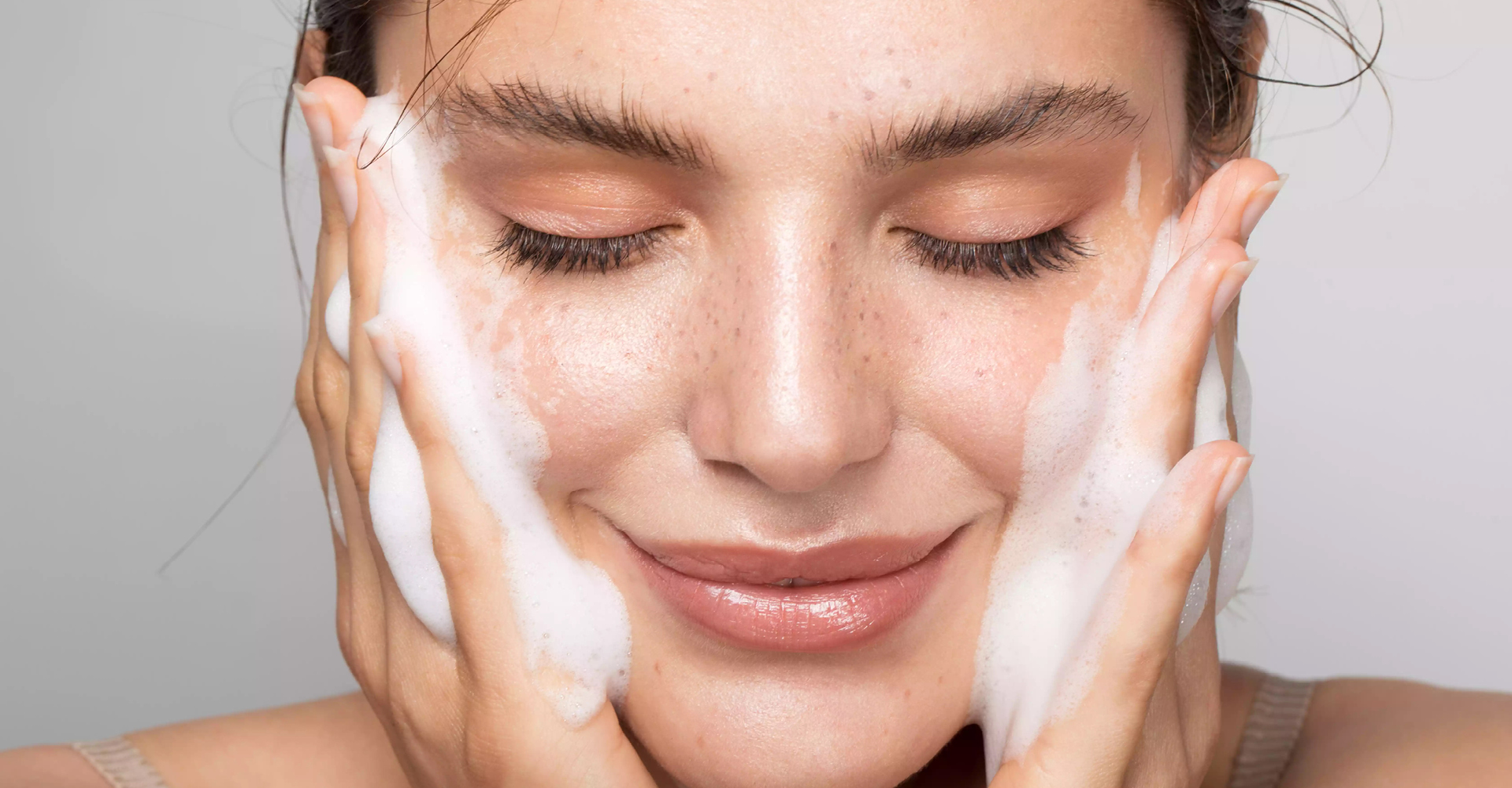 immagine ragazza che sorride, con due mani con mousse per pulirsi il viso. Si tocca le guance